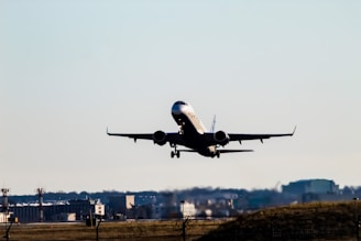 A commercial airplane is taking off from an airport, with its landing gear still extended. The background shows airport infrastructure including terminals and control towers, set against a clear blue sky.