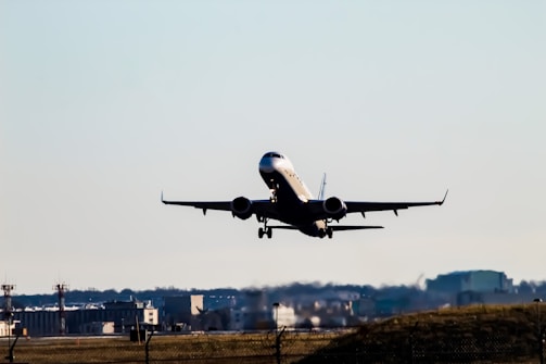 A commercial airplane is taking off from an airport, with its landing gear still extended. The background shows airport infrastructure including terminals and control towers, set against a clear blue sky.