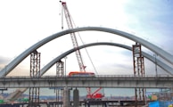 A large red crane is situated beside a bridge under construction, which has two prominent arch structures. A colorful bus crosses the bridge while the incomplete sections are supported by tall metal pillars. The background shows a city skyline with a cloudy sky.