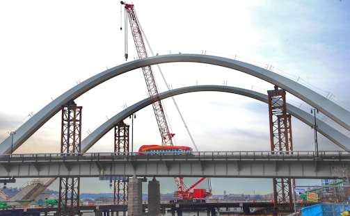 A large red crane is situated beside a bridge under construction, which has two prominent arch structures. A colorful bus crosses the bridge while the incomplete sections are supported by tall metal pillars. The background shows a city skyline with a cloudy sky.