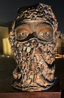 A large sculpture composed of an assortment of objects forming a human face. The eyes are prominent, and the structure appears to be made from various metallic components including pots, pipes, and gears. The lighting enhances the textures and shadows, creating a striking visual effect.