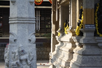 A stone pillar with intricate carvings stands prominently in front of a temple structure. The temple walls are adorned with small deity sculptures and decorated with garlands of bright yellow flowers. Sunlight casts shadows on the textured stone surfaces, creating a serene ambiance. Signs in both a local script and English label the deities.