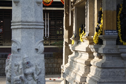 A stone pillar with intricate carvings stands prominently in front of a temple structure. The temple walls are adorned with small deity sculptures and decorated with garlands of bright yellow flowers. Sunlight casts shadows on the textured stone surfaces, creating a serene ambiance. Signs in both a local script and English label the deities.