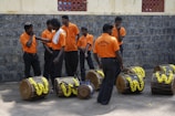 A group of men wearing matching orange shirts and dark pants stands beside traditional drums adorned with yellow flowers. They appear to be engaged in conversation or preparing for an event, with a stone wall and part of a building visible in the background.