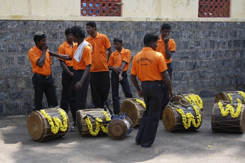 A group of men wearing matching orange shirts and dark pants stands beside traditional drums adorned with yellow flowers. They appear to be engaged in conversation or preparing for an event, with a stone wall and part of a building visible in the background.
