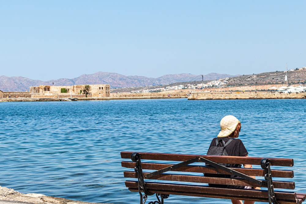 man in brown jacket sitting on brown wooden bench near body of water during daytime