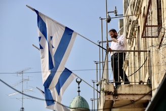 A man is standing on a balcony holding a large flag with blue and white stripes and a Star of David. He appears to be adjusting something, possibly a flagpole. The building is constructed with stone and there are various metal fixtures surrounding the area.