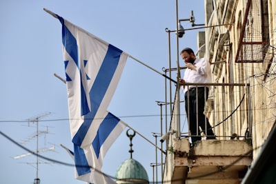 A man is standing on a balcony holding a large flag with blue and white stripes and a Star of David. He appears to be adjusting something, possibly a flagpole. The building is constructed with stone and there are various metal fixtures surrounding the area.