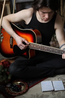 A person wearing a black tank top is seated on the floor, playing an acoustic guitar. The guitar has a sunburst finish and is held with one hand strumming. Nearby, there is a plate with grapes and a notebook with handwritten notes. Greenery is visible in the corner, suggesting a cozy, indoor setting.