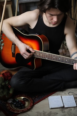 A person wearing a black tank top is seated on the floor, playing an acoustic guitar. The guitar has a sunburst finish and is held with one hand strumming. Nearby, there is a plate with grapes and a notebook with handwritten notes. Greenery is visible in the corner, suggesting a cozy, indoor setting.