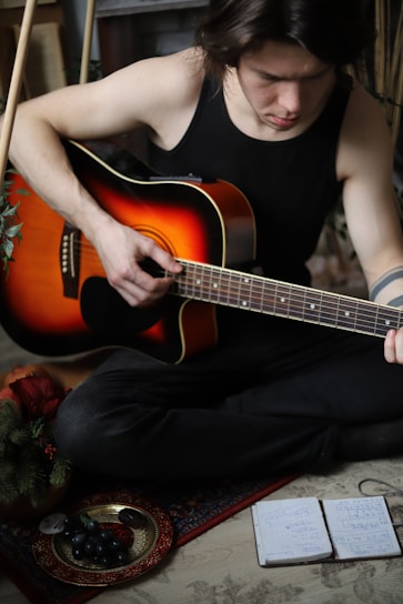 A person wearing a black tank top is seated on the floor, playing an acoustic guitar. The guitar has a sunburst finish and is held with one hand strumming. Nearby, there is a plate with grapes and a notebook with handwritten notes. Greenery is visible in the corner, suggesting a cozy, indoor setting.