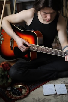 A person wearing a black tank top is seated on the floor, playing an acoustic guitar. The guitar has a sunburst finish and is held with one hand strumming. Nearby, there is a plate with grapes and a notebook with handwritten notes. Greenery is visible in the corner, suggesting a cozy, indoor setting.