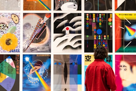 A person wearing a red jacket observes a large display of colorful and diverse artwork, featuring abstract patterns, vibrant geometric designs, and cultural motifs. The artwork is arranged in a grid on a wall, showcasing a variety of themes and styles.
