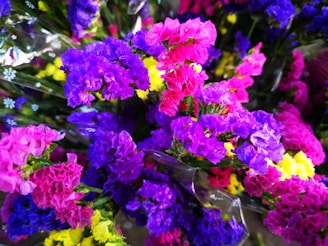 A vibrant assortment of colorful flowers, featuring hues of purple, pink, yellow, and blue. The flowers appear to be wrapped in a translucent plastic material, with some smaller white floral patterns visible.