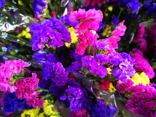 A vibrant assortment of colorful flowers, featuring hues of purple, pink, yellow, and blue. The flowers appear to be wrapped in a translucent plastic material, with some smaller white floral patterns visible.