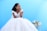 A bride in an elegant white wedding dress with intricate lace detailing sits on a decorative chair against a bright blue background. To her right, there is a beautiful floral arrangement with white calla lilies and small red flowers. Her hair is styled in loose curls, and she appears to be looking thoughtfully toward the distance with a gentle smile.