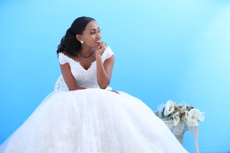 A bride in an elegant white wedding dress with intricate lace detailing sits on a decorative chair against a bright blue background. To her right, there is a beautiful floral arrangement with white calla lilies and small red flowers. Her hair is styled in loose curls, and she appears to be looking thoughtfully toward the distance with a gentle smile.