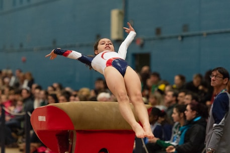 A gymnast is in mid-air performing a vault maneuver with her arms extended. She wears a white leotard with red and blue designs. In the background, a crowd of spectators is watching the performance in an indoor gymnasium.