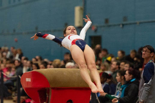 A gymnast is in mid-air performing a vault maneuver with her arms extended. She wears a white leotard with red and blue designs. In the background, a crowd of spectators is watching the performance in an indoor gymnasium.