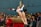 A gymnast is in mid-air performing a vault maneuver with her arms extended. She wears a white leotard with red and blue designs. In the background, a crowd of spectators is watching the performance in an indoor gymnasium.