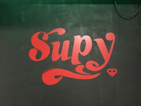 A stylized red text reading 'Supy' is displayed on a dark green, possibly chalkboard-like, background. To the bottom left of the text, there appears to be a faint additional text or marking. A circular and ornate design element is seen below the main text.