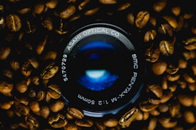 A camera lens with branding 'ASAHI OPTICAL CO.' and 'SMC PENTAX' surrounded by roasted coffee beans.