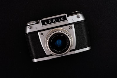 A vintage Exa 1a camera with a metallic silver and black exterior, featuring a prominent lens and various dials and buttons for manual adjustments, placed against a dark background.