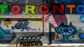 Graffiti art on a brick wall featuring the word 'Toronto' in large, colorful letters. Below, various cartoon-style characters are depicted, including a large cat with prominent eyes, a squirrel with hockey gear, and other playful figures. Gas meters and pipes are present at the bottom of the mural.