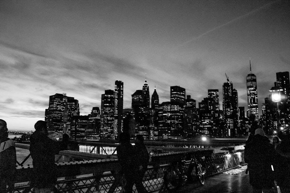 grayscale photo of man standing on bridge near city buildings
