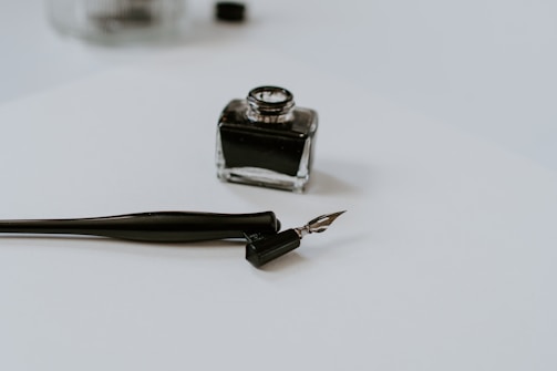 A close-up of a calligraphy pen and an ink bottle on a plain white surface. The pen is uncapped and the nib is resting on the paper. The ink bottle is small and square with a dark liquid inside.