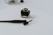 A close-up of a calligraphy pen and an ink bottle on a plain white surface. The pen is uncapped and the nib is resting on the paper. The ink bottle is small and square with a dark liquid inside.