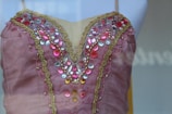 A detailed view of a pink garment adorned with intricate lace and an array of colorful gemstones. The gems include shades of red, pink, and silver, arranged symmetrically along the neckline, with a gold trim outlining the edges.