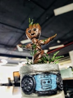 A small, detailed statuette of a character with a cute face, resembling a baby tree. The figurine is crafted with brown and green elements, indicating bark and leaves. It stands on a pot with an abstract design and is placed in a modern office setting.