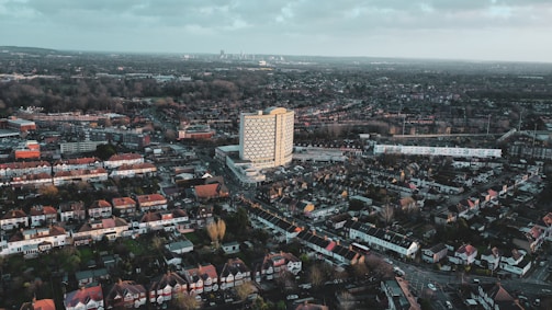 An aerial view of a cityscape featuring a prominent high-rise building surrounded by densely packed residential areas with rows of houses. The scene extends to the horizon, showing a blend of urban and suburban environments under a partly cloudy sky.
