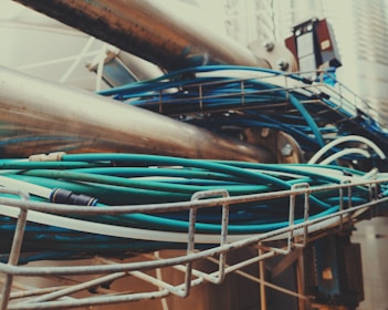 A complex arrangement of metal pipes and teal-colored cables, intertwined and secured in a metal framework. The setting appears industrial, with pipes and cables extending in various directions.