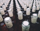 A large collection of glass bottles arranged in rows, each with a beige paper label around the neck featuring a stylized design and text. The tops of the bottles are sealed with metallic caps.