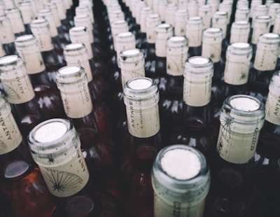 A large collection of glass bottles arranged in rows, each with a beige paper label around the neck featuring a stylized design and text. The tops of the bottles are sealed with metallic caps.