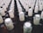 A large collection of glass bottles arranged in rows, each with a beige paper label around the neck featuring a stylized design and text. The tops of the bottles are sealed with metallic caps.
