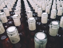 A large collection of glass bottles arranged in rows, each with a beige paper label around the neck featuring a stylized design and text. The tops of the bottles are sealed with metallic caps.