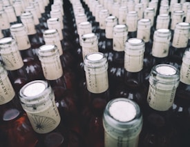 A large collection of glass bottles arranged in rows, each with a beige paper label around the neck featuring a stylized design and text. The tops of the bottles are sealed with metallic caps.