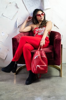 A person wearing a fashionable red outfit and heart-shaped sunglasses sits on a plush red chair. The individual holds a shiny red backpack and has an air of relaxed confidence. The background has paper sheets attached to a wall, adding an artistic flair.