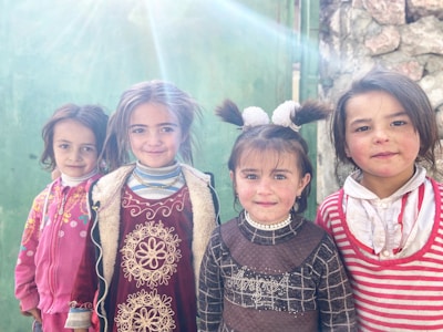 Four young children, dressed colorfully, stand close together with cheerful expressions. Bright light creates a sun flare effect in the top left, and they are surrounded by a textured, green background and a stone wall.