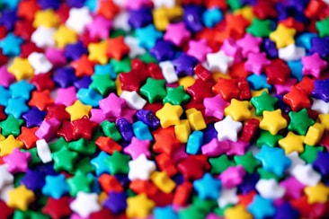 A vibrant collection of tiny, multicolored star-shaped candies scattered across a surface. The stars are in various shades of red, pink, blue, green, yellow, purple, white, and orange, creating a visually striking and cheerful pattern.