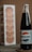 A vintage glass bottle labeled with a retro-style Pepsi-Cola logo is placed on a striped surface. The bottle's contents are dark, suggesting a cola beverage. In the background, there is a box with a column of stacked round crackers, slightly out of focus.