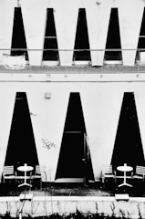 Geometric black and white design features large vertical black triangles on a white wall with two outdoor seating arrangements. Each arrangement includes a round white table with two striped chairs. The minimalistic and modern architecture contrasts sharply with the black shapes, creating a striking visual effect.