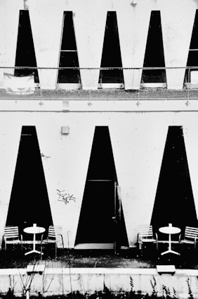 Geometric black and white design features large vertical black triangles on a white wall with two outdoor seating arrangements. Each arrangement includes a round white table with two striped chairs. The minimalistic and modern architecture contrasts sharply with the black shapes, creating a striking visual effect.