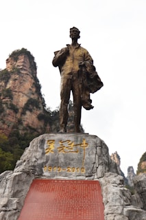 A bronze statue of a man holding a book and a coat, standing atop a rocky pedestal inscribed with golden characters and the dates 1919-2010. In the background, tall, rocky cliffs covered with some vegetation are visible under a partly cloudy sky.