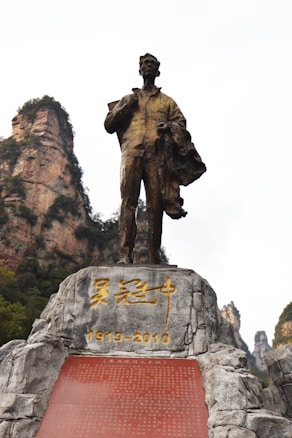A bronze statue of a man holding a book and a coat, standing atop a rocky pedestal inscribed with golden characters and the dates 1919-2010. In the background, tall, rocky cliffs covered with some vegetation are visible under a partly cloudy sky.