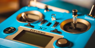 A close-up of a blue remote control device featuring several buttons, switches, and joysticks. The surface is smooth with metallic accents, and the device is labeled 'Taranis Q X7'. The lighting highlights the texture and details of the controls.