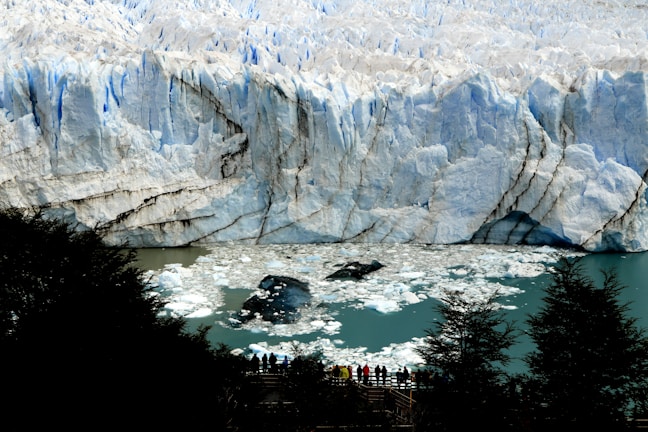 A massive glacier with towering icy ridges dominates the background, revealing a textured surface marked by cracks and crevices. In the foreground, a group of people stand on a viewing platform, silhouetted against the vibrant turquoise water filled with floating ice chunks.